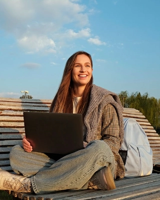Eine junge Frau sitzt am einen sonnigen Tag auf einer Bank und arbeitet am Laptop.